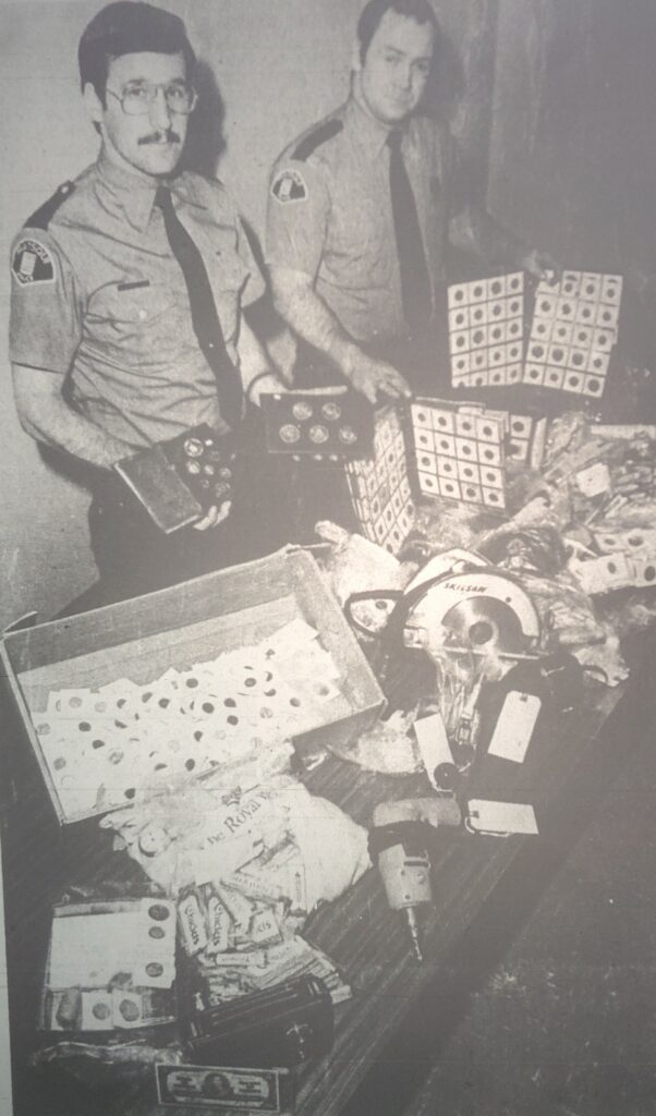 Two officers pose with stolen goods, including power tools, money, and collectable coins.