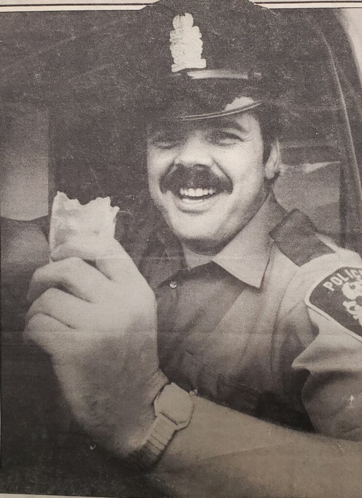 photo of a cop smiling at the camera with a sandwich in his hand.