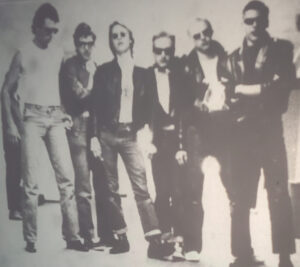 A group of six teenage boys pose in leather jackets and sunglasses giving off a 1960's greaser look